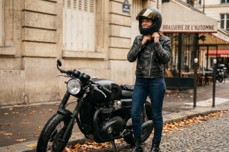 Femme motarde en veste en cuir noir ajustant son casque intégral matte devant une moto café racer sur des pavés parisiens